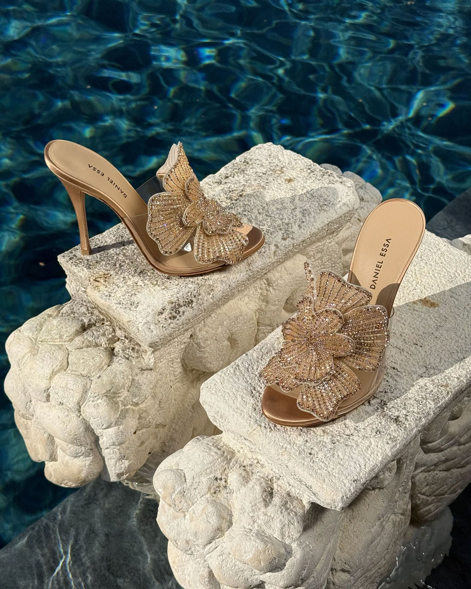 Pair of Daniel Essa high-heeled sandals featuring artisanal floral embellishments, displayed on a stone ledge in an elegant poolside setting