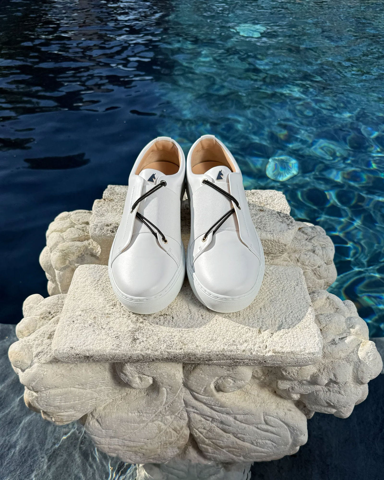 Daniel Essa Toi&Moi sneakers in white leather with interchangeable black elastic laces, displayed on a stone surface by a poolside